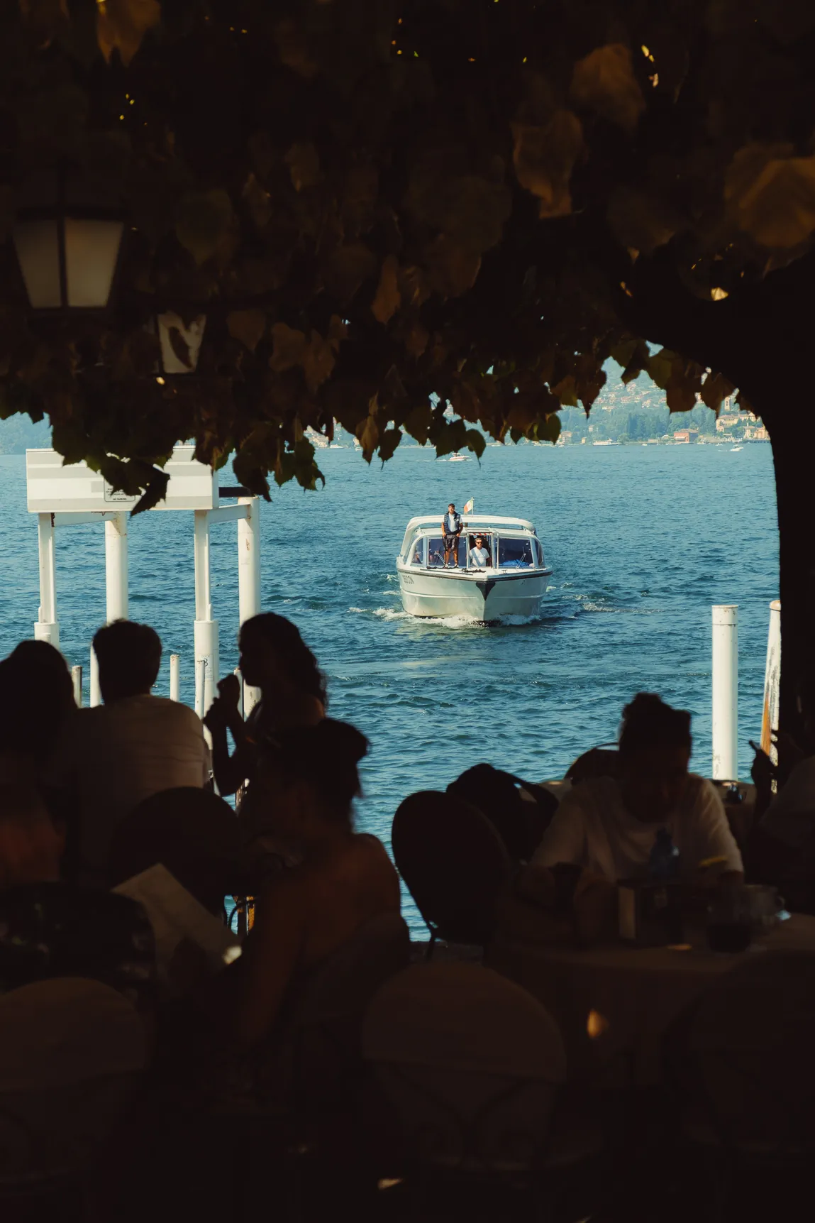 Een watertaxi vaart aan, ingekaderd door bladeren en silhouetten op een terras
